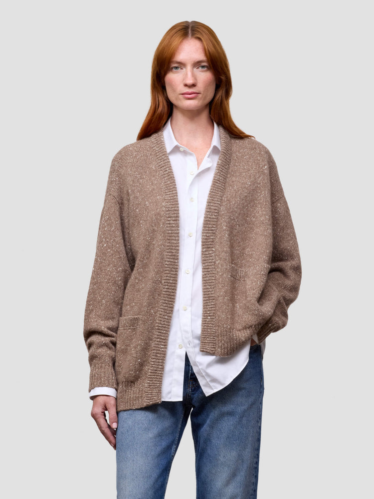 Woman wearing a brown cashmere cardigan over a white shirt and blue jeans on a light gray background