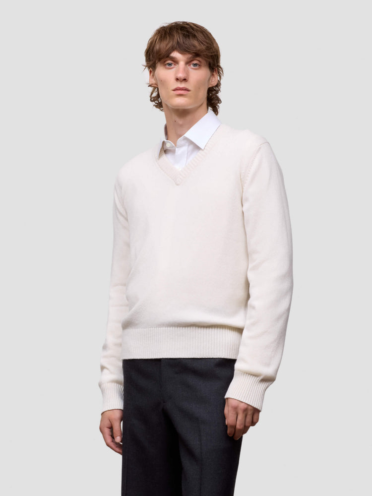 Man wearing a white cashmere v-neck sweater over a collared shirt with dark pants on a light gray background