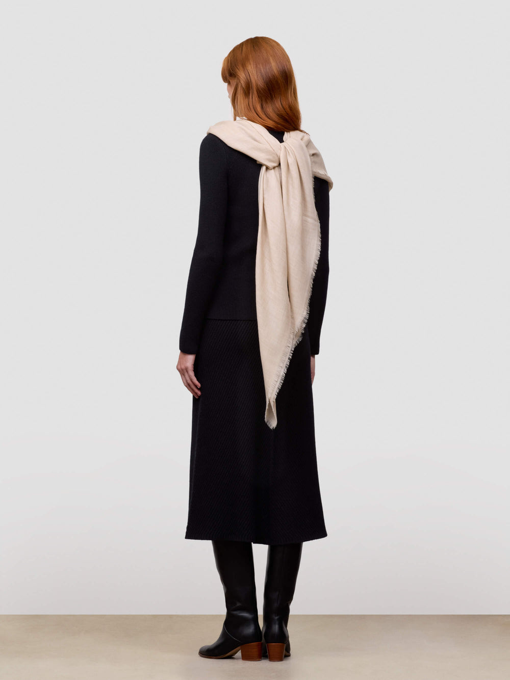 Woman wearing a black cashmere sweater and skirt with beige cashmere scarf tied behind back against a white background