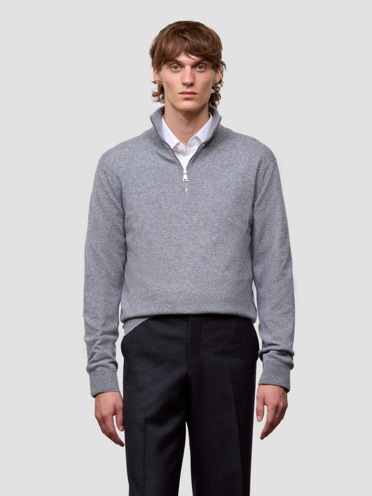 Man wearing a gray half zip sweater over a white shirt with black pants on a light gray background