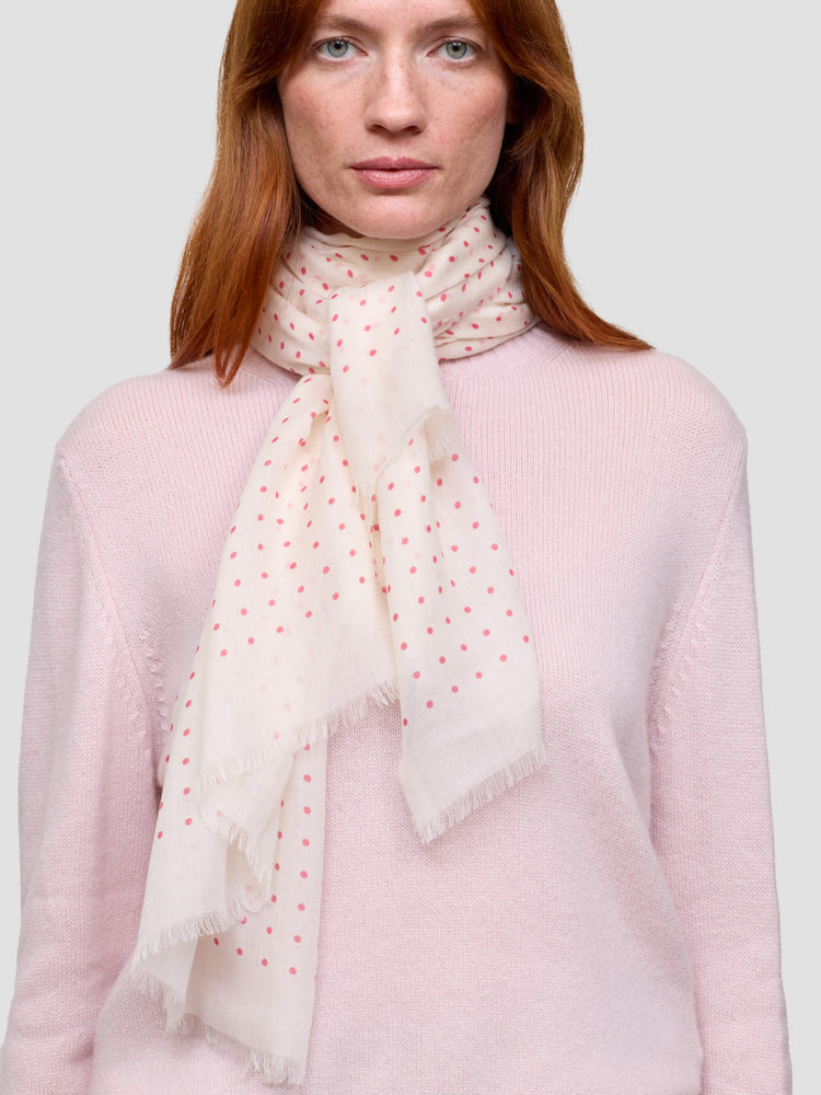 Woman wearing a light pink sweater and a white scarf with red polka dots on a plain background