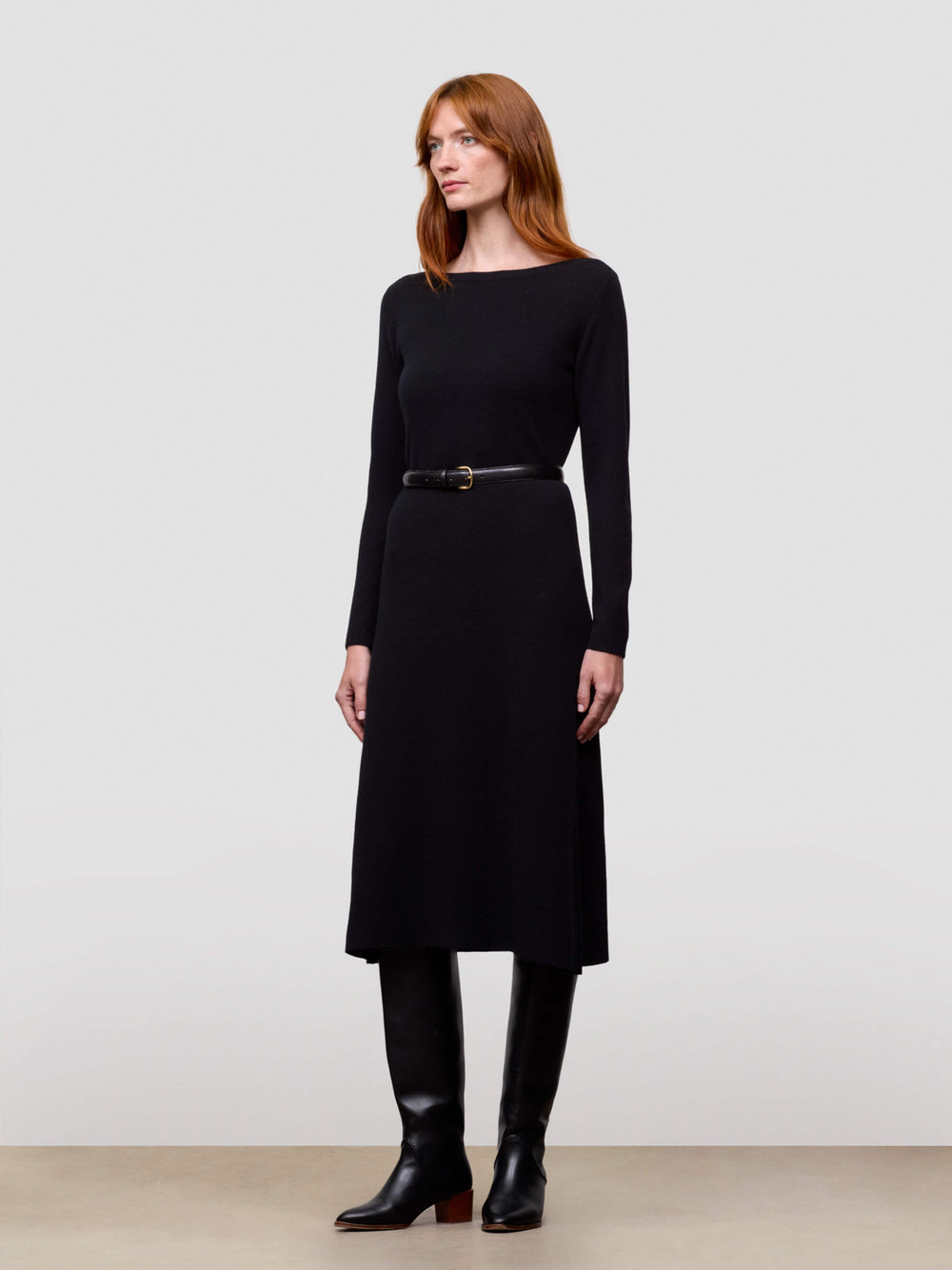 Woman wearing a black dress with a belt and black boots on a plain background