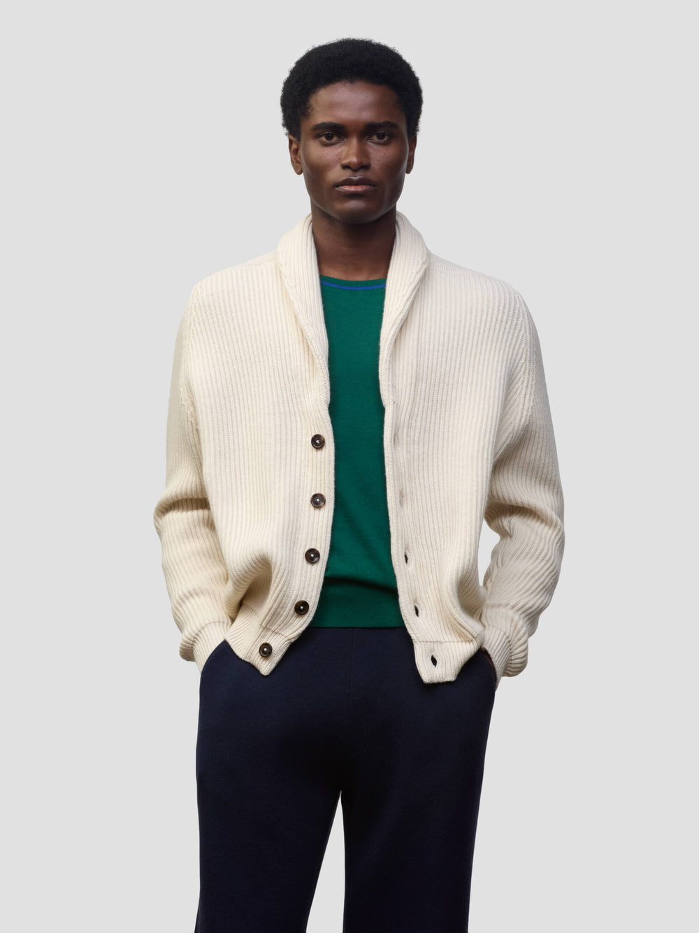man wearing a natural colour ribbed cashmere cardigan over a green tshirt and navy trousers on a grey background