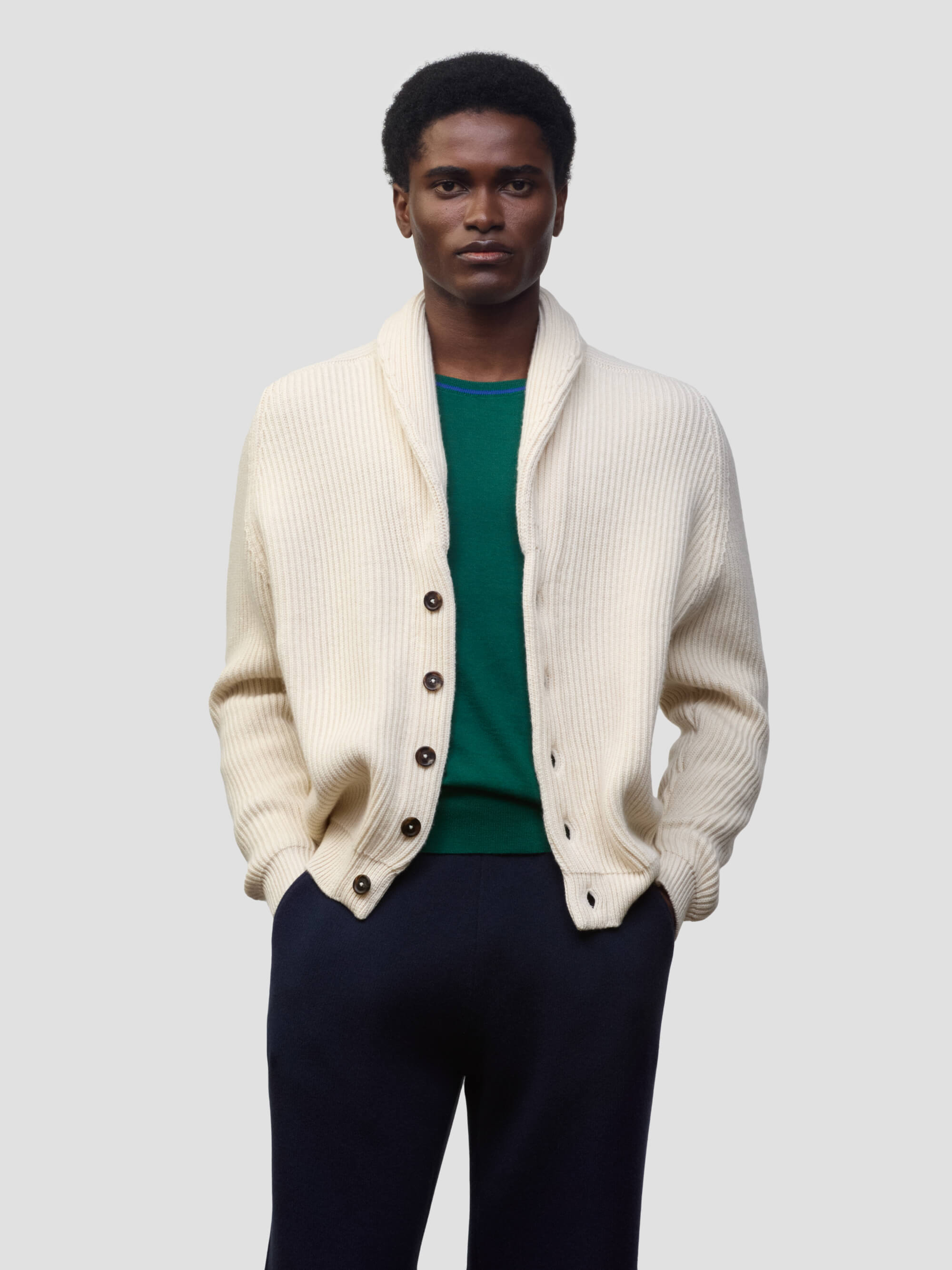 man wearing a natural colour ribbed cashmere cardigan over a green tshirt and navy trousers on a grey background
