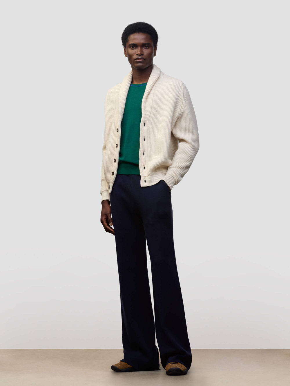 man wearing a natural colour ribbed cashmere cardigan over a green tshirt and navy trousers on a grey background