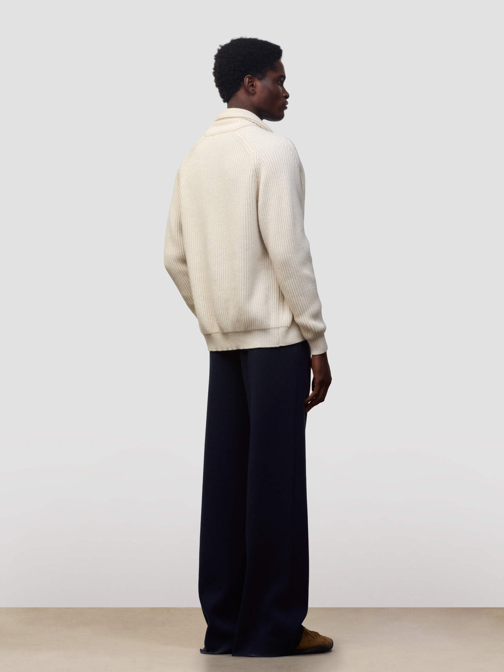 man wearing a natural colour ribbed cashmere cardigan over a green tshirt and navy trousers on a grey background