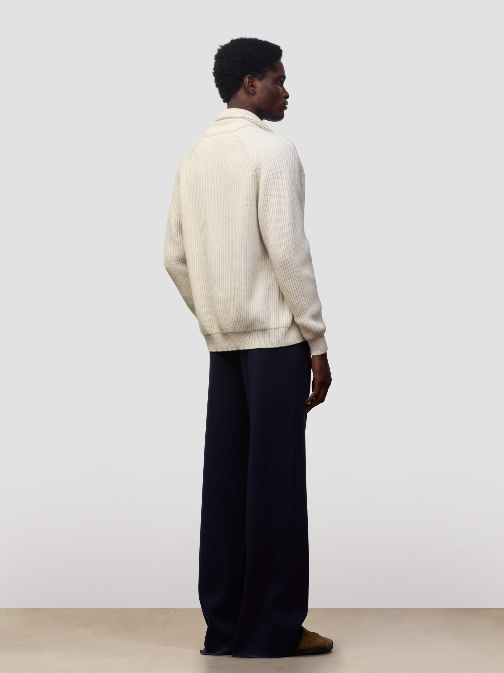 man wearing a natural colour ribbed cashmere cardigan over a green tshirt and navy trousers on a grey background