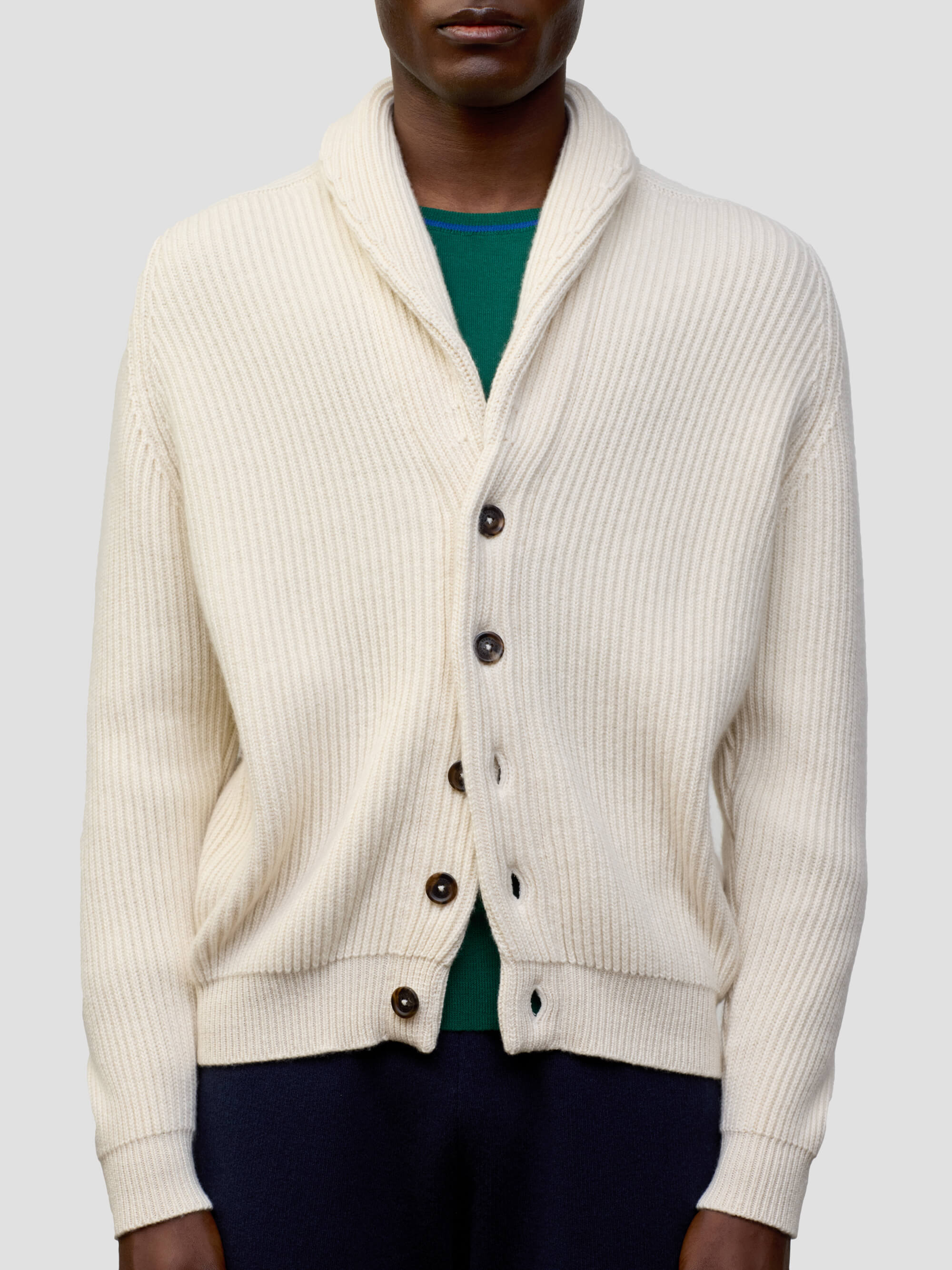 man wearing a natural colour ribbed cashmere cardigan over a green tshirt and navy trousers on a grey background