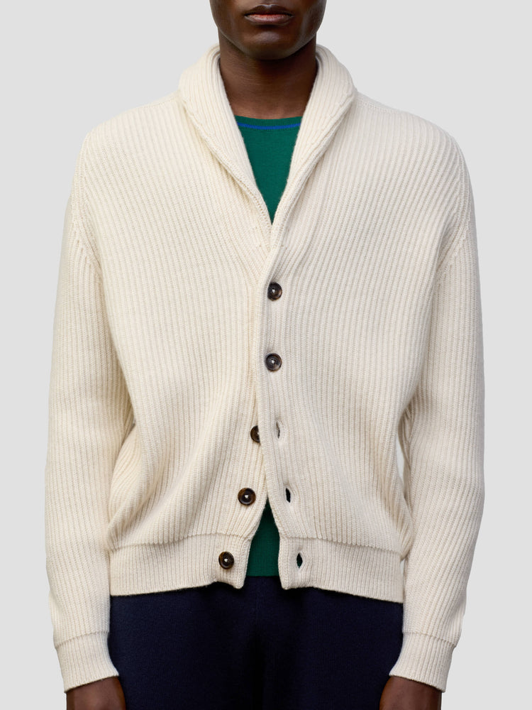 man wearing a natural colour ribbed cashmere cardigan over a green tshirt and navy trousers on a grey background