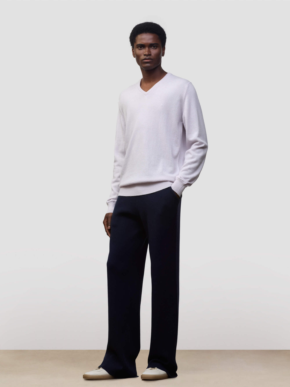 man wearing a light pink cashmere v-neck sweater and navy trousers on a grey background