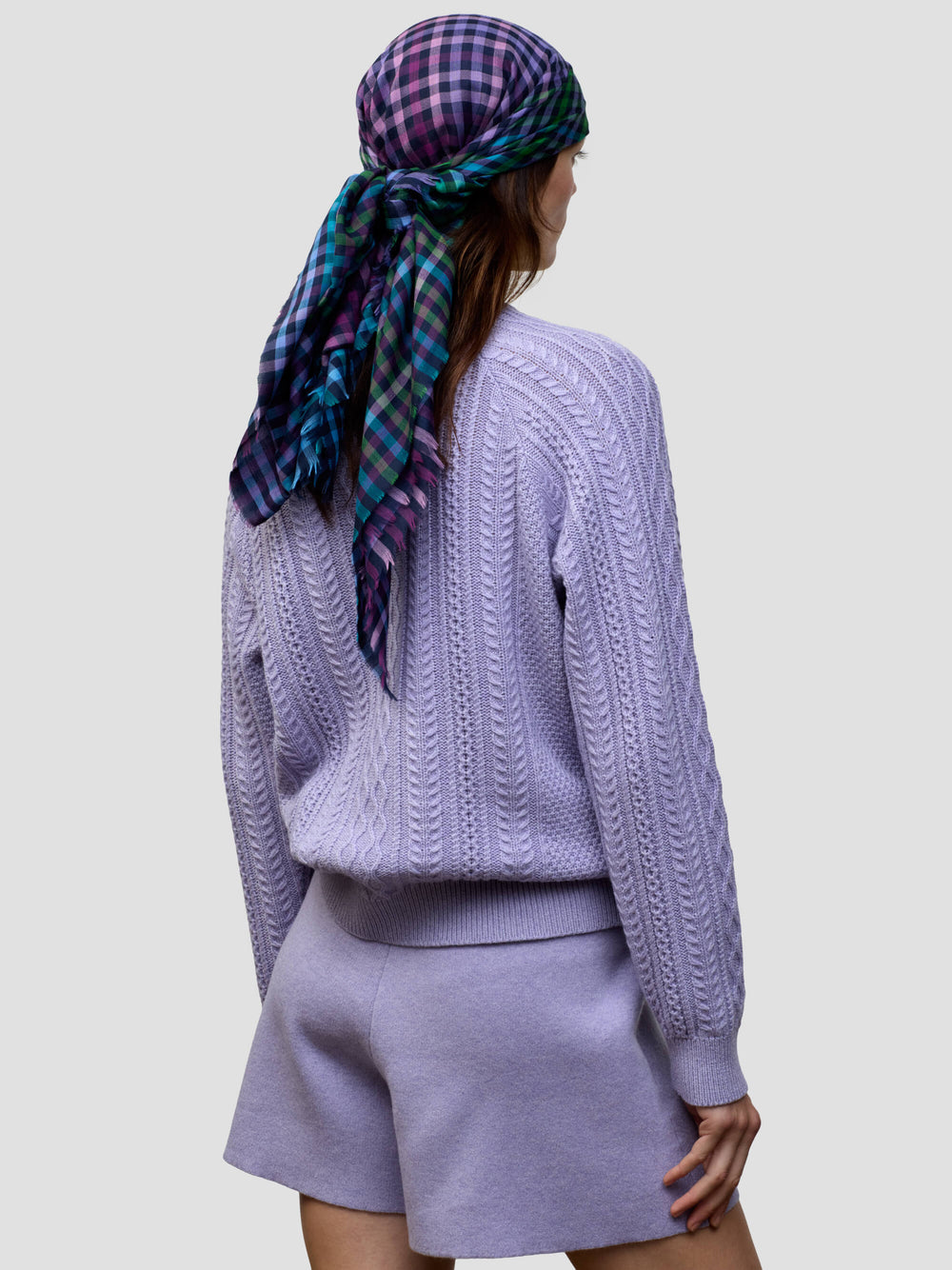 woman wearing a cable knit purple sweater and matching shorts with a check purple, green and blue scarf tied around head on a grey background
