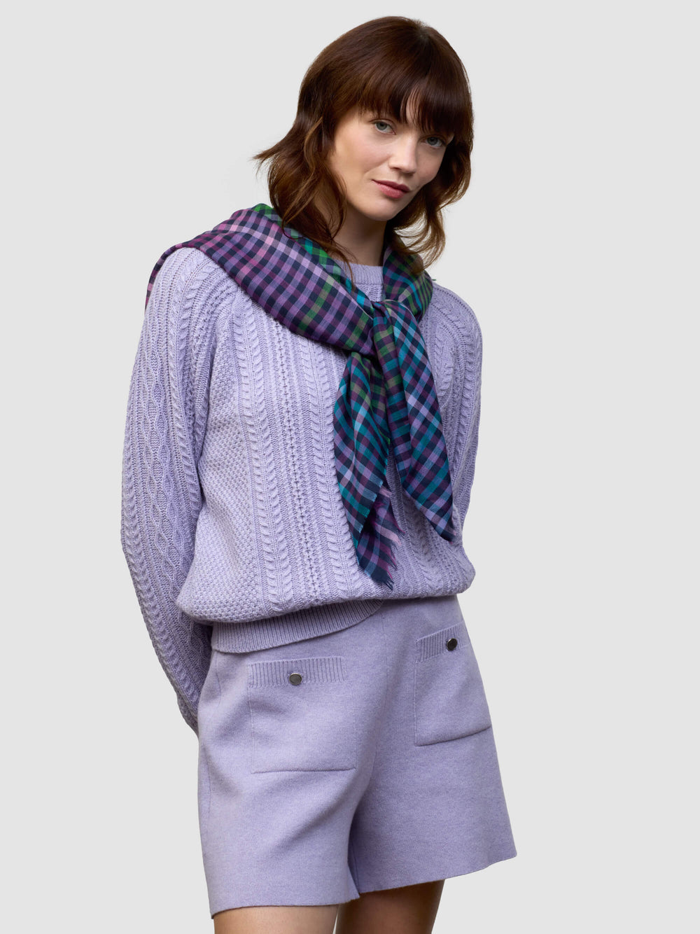 woman wearing a cable knit purple sweater and matching shorts with a check purple, green and blue scarf on a grey background
