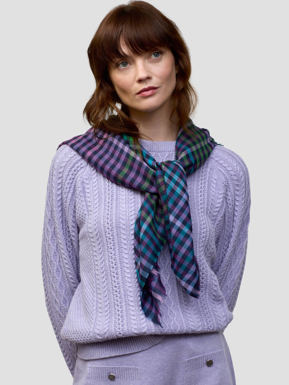 woman wearing a cable knit purple sweater and matching shorts with a check purple, green and blue scarf on a grey background