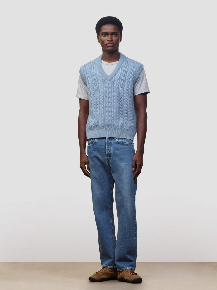 man wearing an arran cable knit blue vest over a grey tshirt and jeans on a grey background
