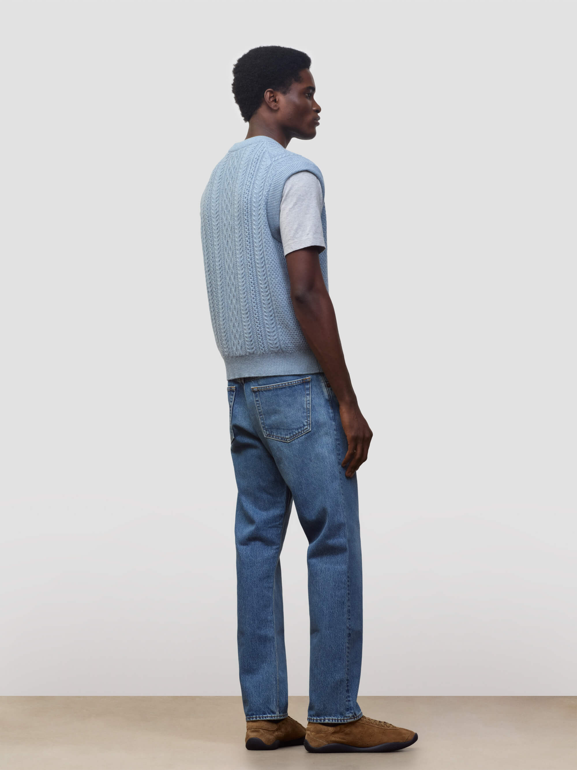 man wearing an arran cable knit blue vest over a grey tshirt and jeans on a grey background