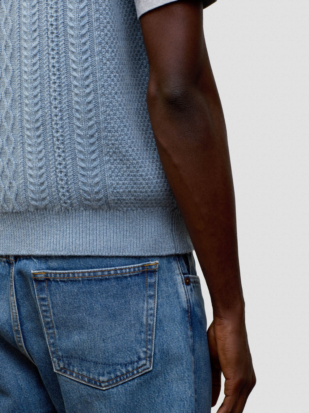 man wearing an arran cable knit blue vest and jeans on a grey background