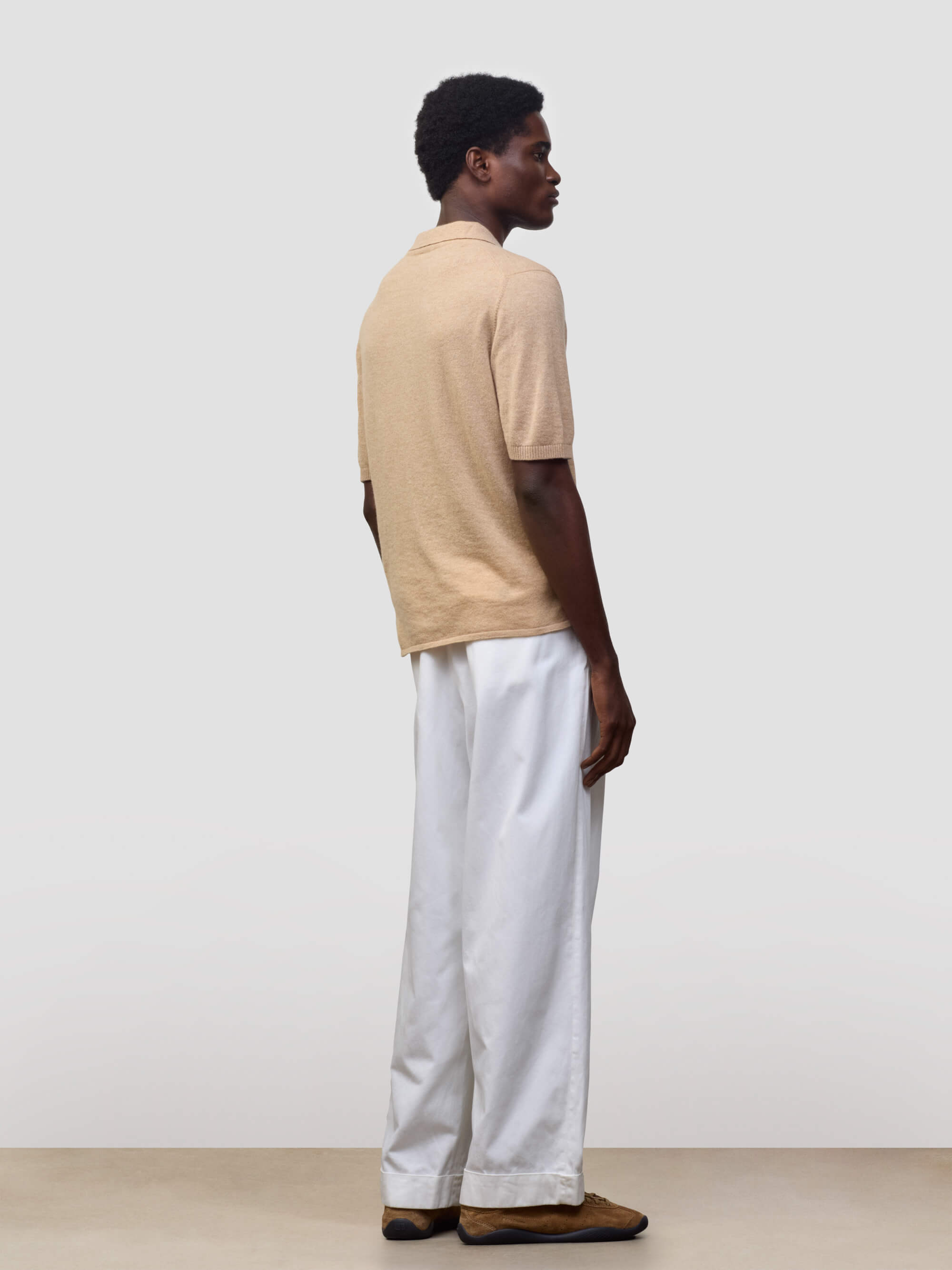 man wearing beige cashmere polo and white trousers on a grey background