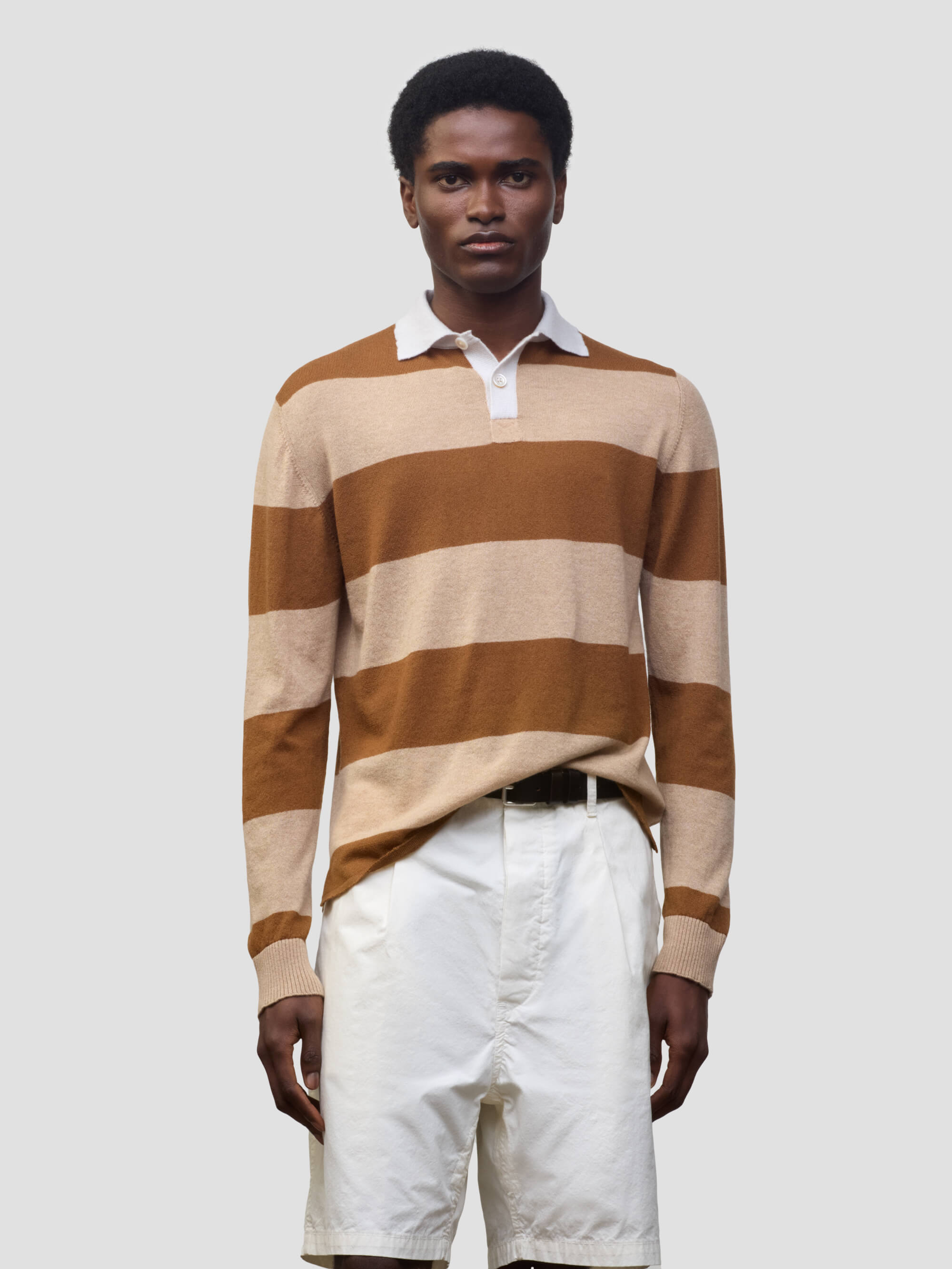 man wearing a striped brown and beige cashmere rugby shirt with white shorts on a grey background