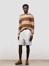 man wearing a striped brown and beige cashmere rugby shirt with white shorts on a grey background