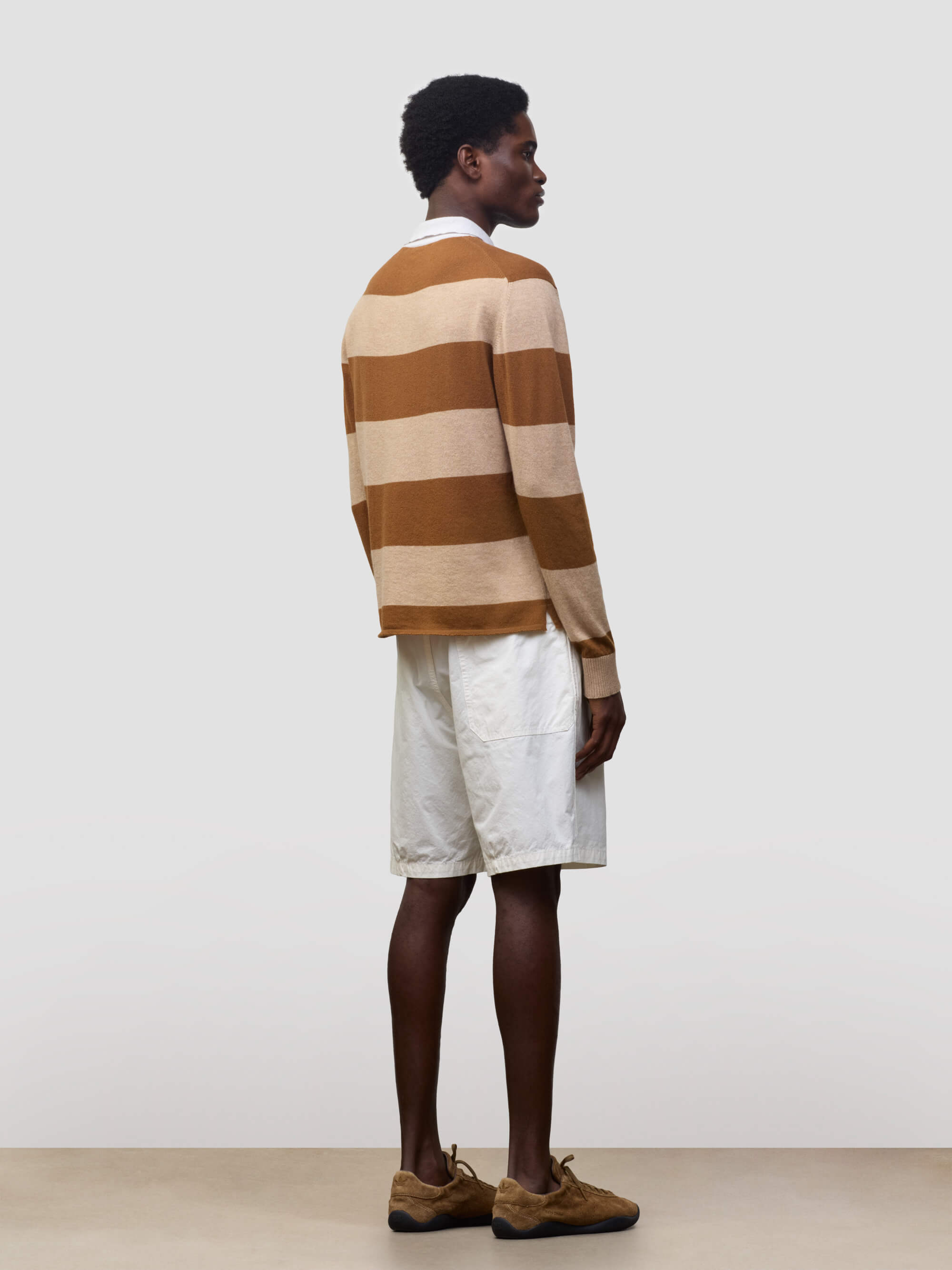 man wearing a striped brown and beige cashmere rugby shirt with white shorts on a grey background
