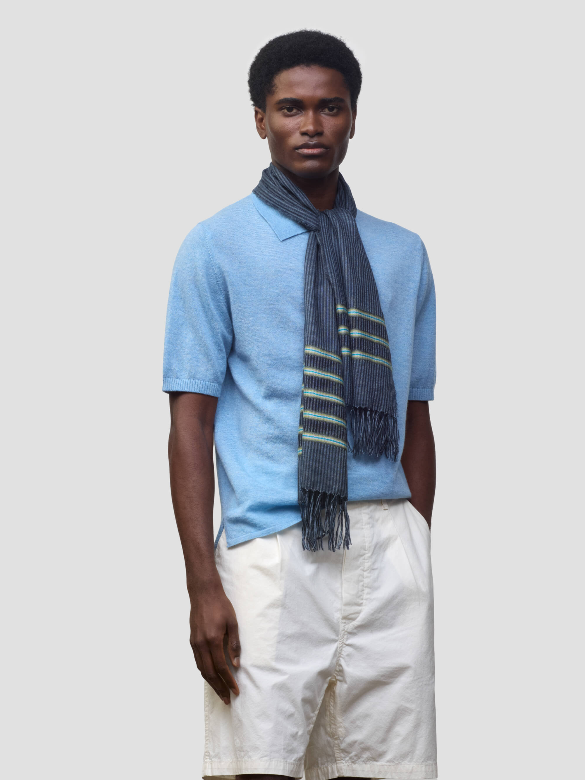 man wearing a light blue cashmere poloshirt with white shorts and navy scarf with green stripes on a grey background