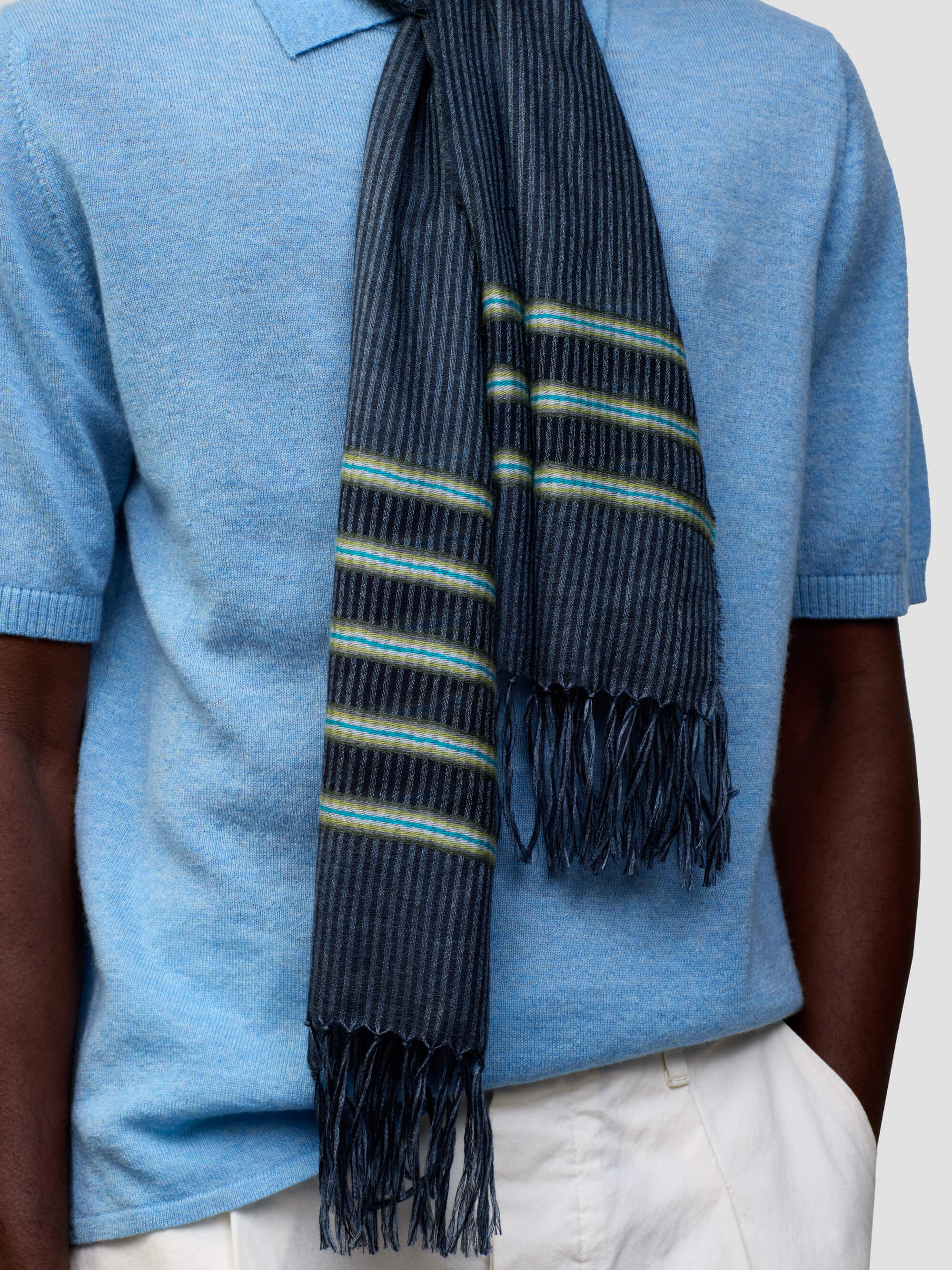 man wearing a light blue cashmere poloshirt with white shorts and navy scarf with green stripes on a grey background