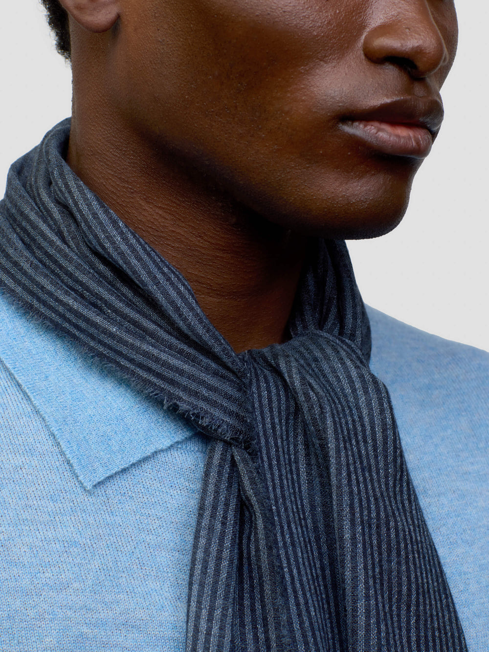 close up of a man wearing a light blue cashmere poloshirt navy scarf stripes on a grey background