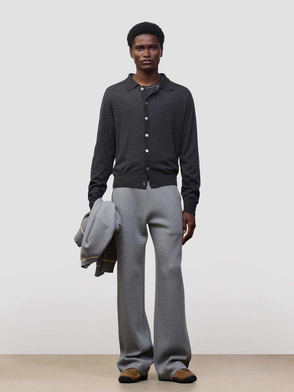Person wearing a dark gray button-up cashmere cardigan over a gray sweater with light gray pants and holding a grey jacket on a white background