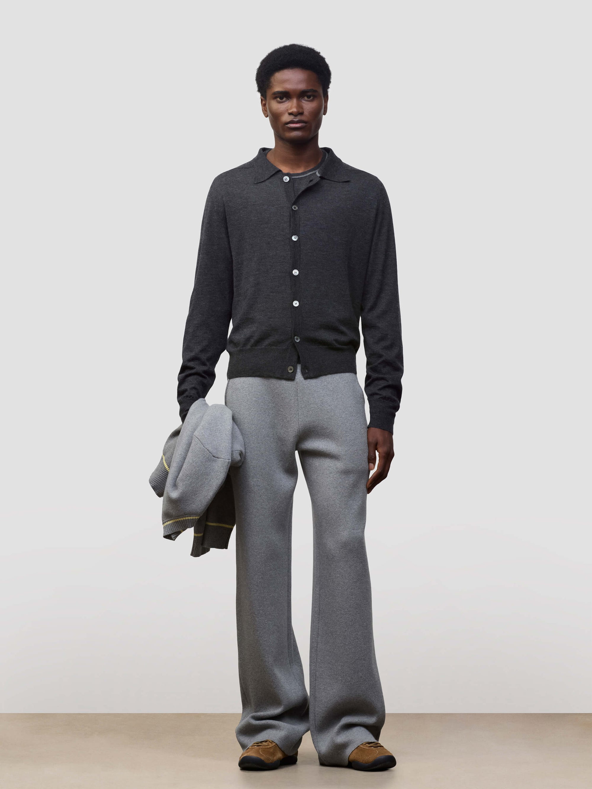 Person wearing a dark gray button-up cashmere cardigan over a gray sweater with light gray pants and holding a grey jacket on a white background