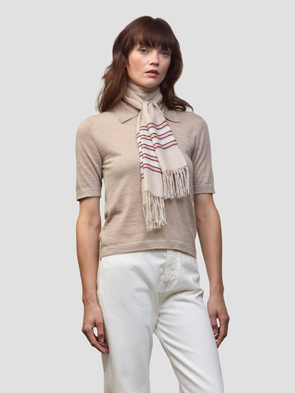 woman wearing a beige cashmere poloshirt and white trousers with beige and pink striped scarf on a grey background