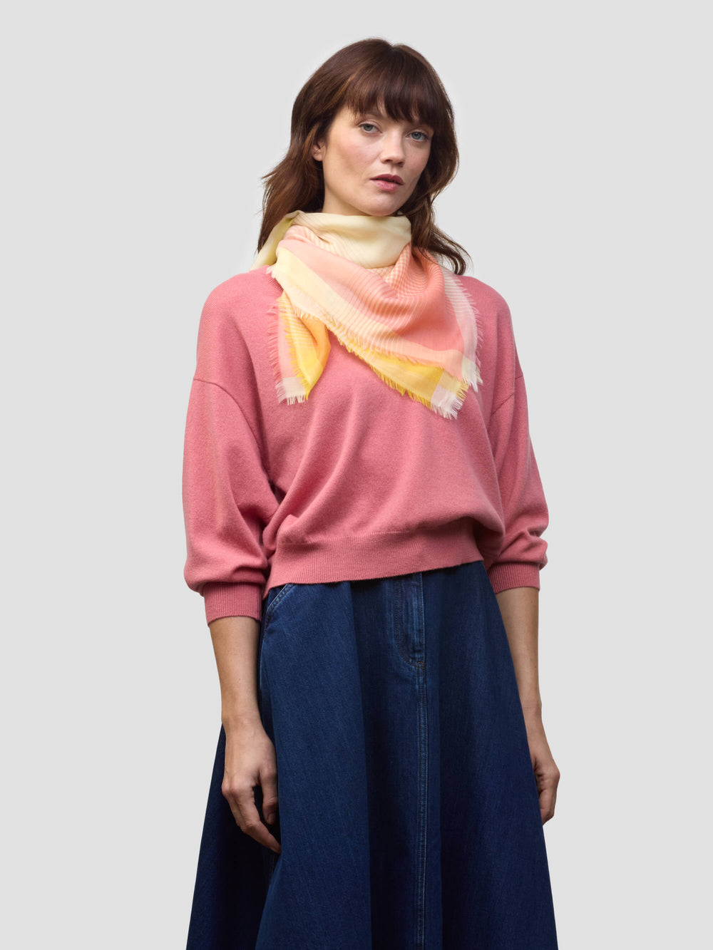woman wearing a pink cashmere sweater with yellow and orange patterned scarf and denim skirt on a grey background