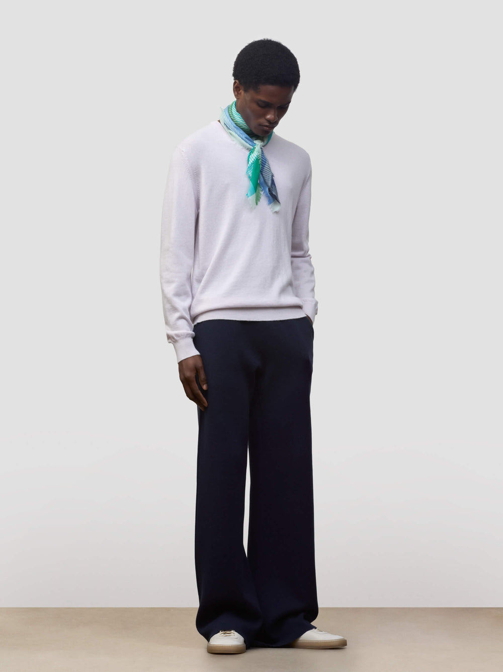 man wearing a lilac cashmere sweater with green and blue patterned neck scarf and navy trousers on a grey background