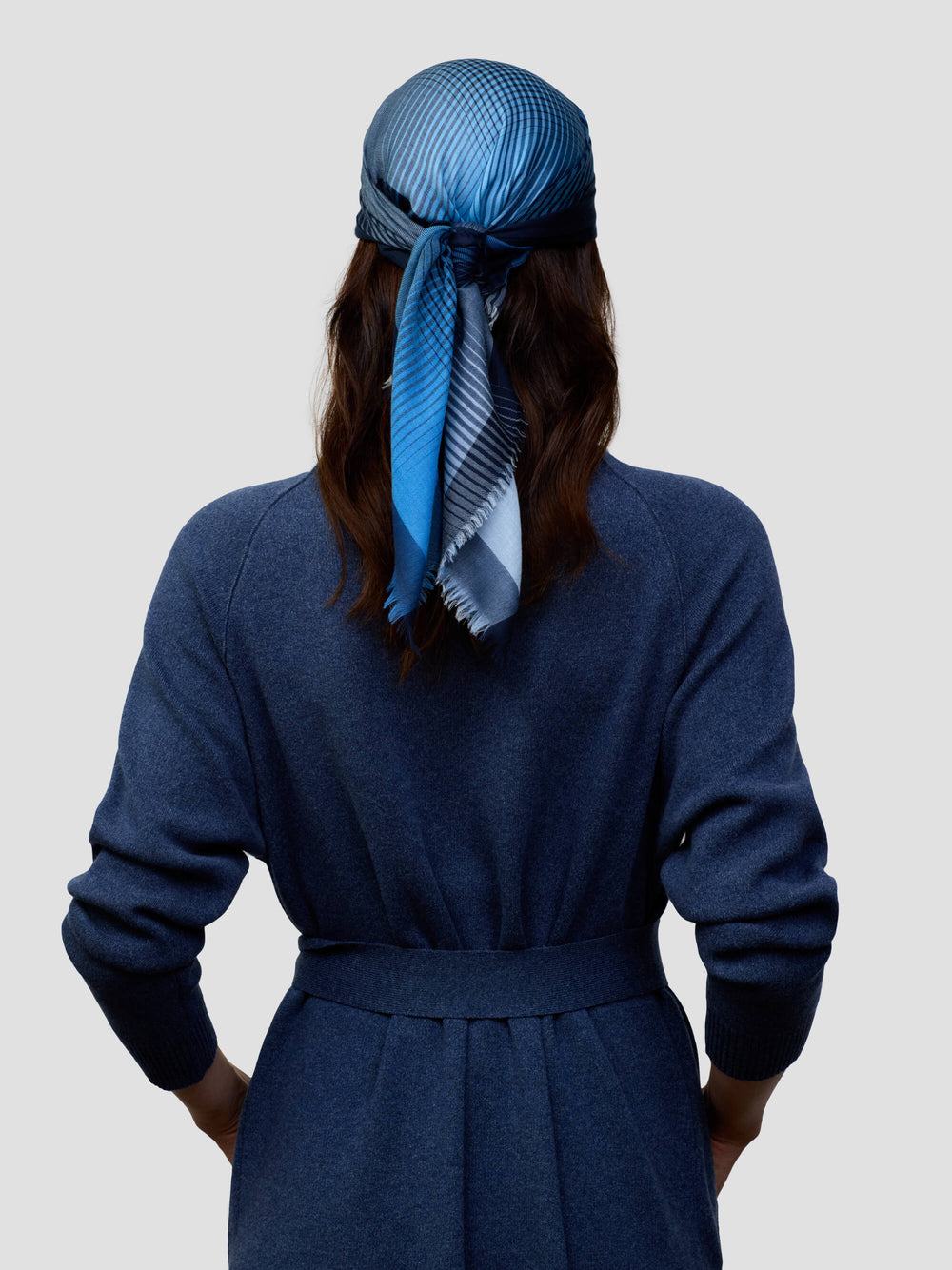 woman wearing a long navy cashmere coat and matching navy and blue patterned scarf tied around head on a grey background