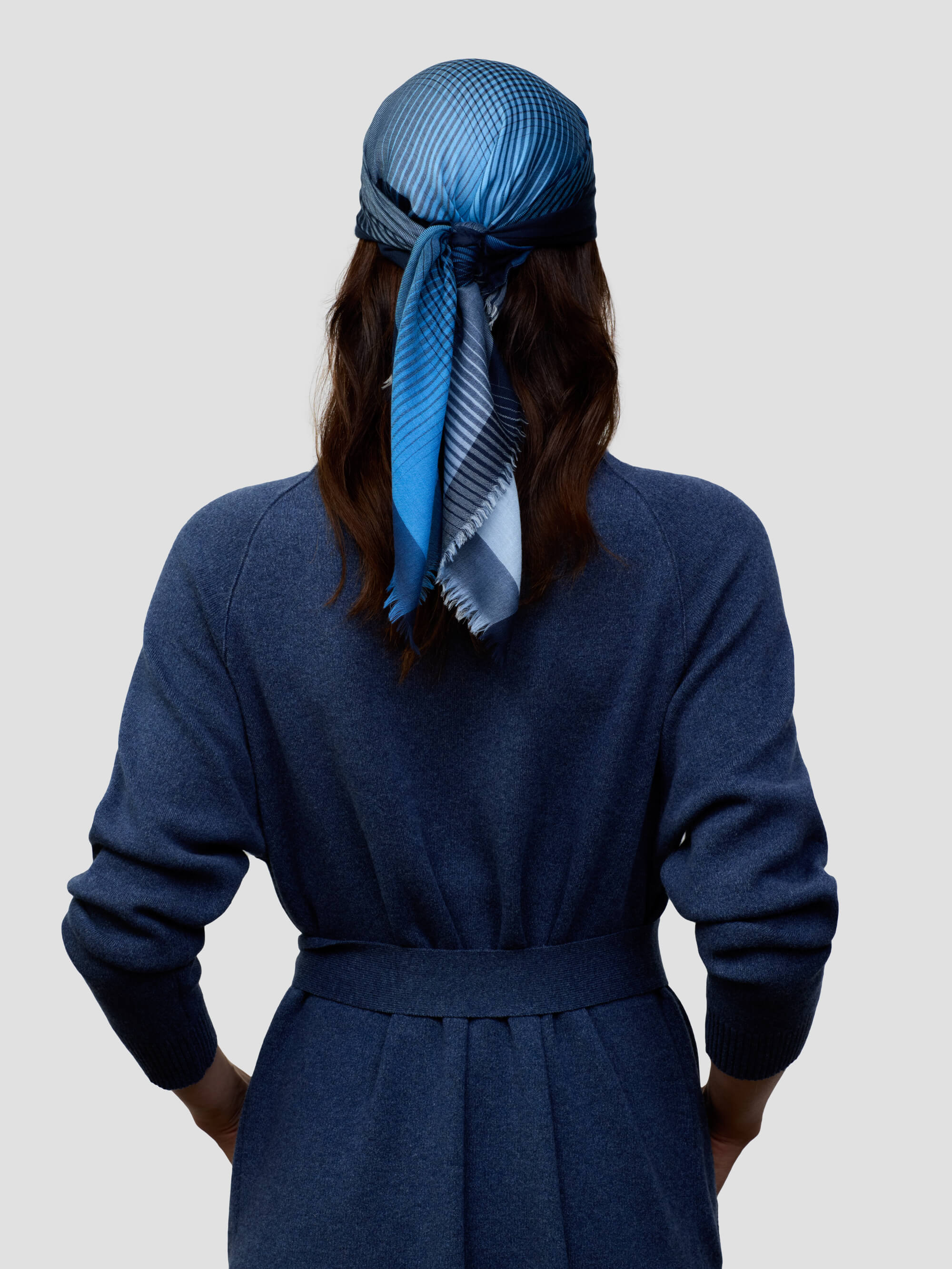 woman wearing a long navy cashmere coat and matching navy and blue patterned scarf tied around head on a grey background