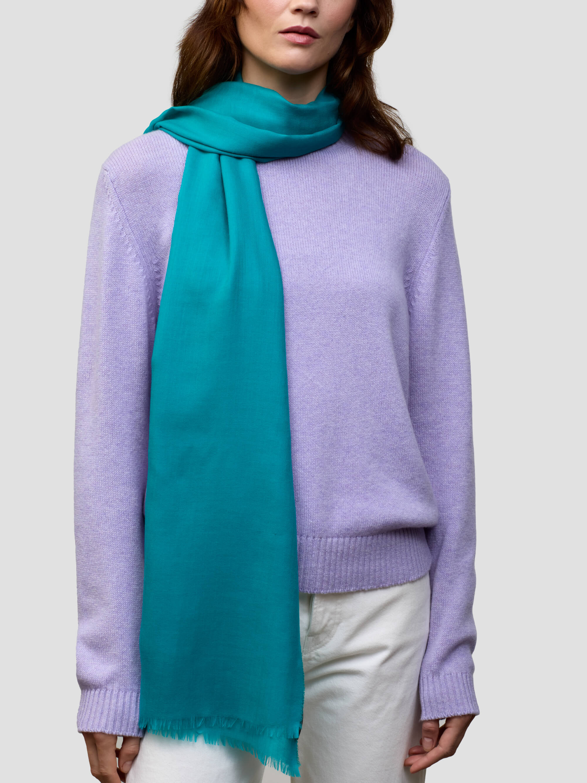 women wearing a lilac cashmere sweater and teal scarf with white trousers on a grey background