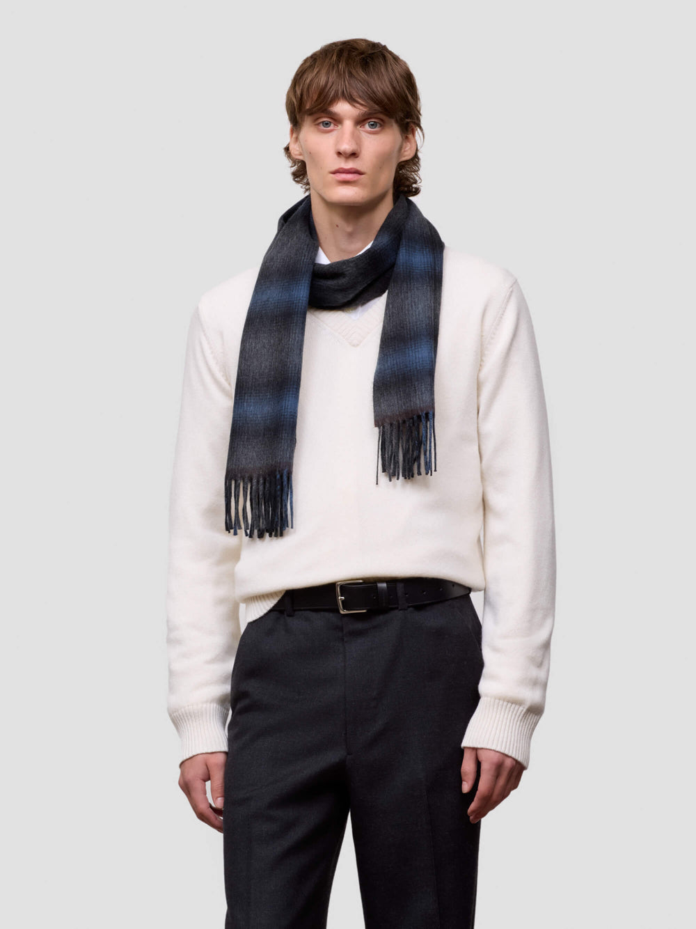 Man wearing a white cashmere sweater and black pants with a blue tartan cashmere scarf on a light gray background