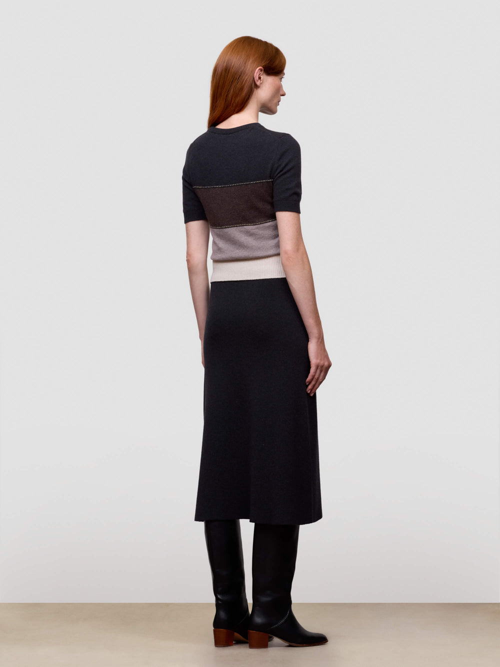 Woman wearing a black, brown, and beige color-blocked cashmere t-shirt on a light gray background and black cashmere dress with boots