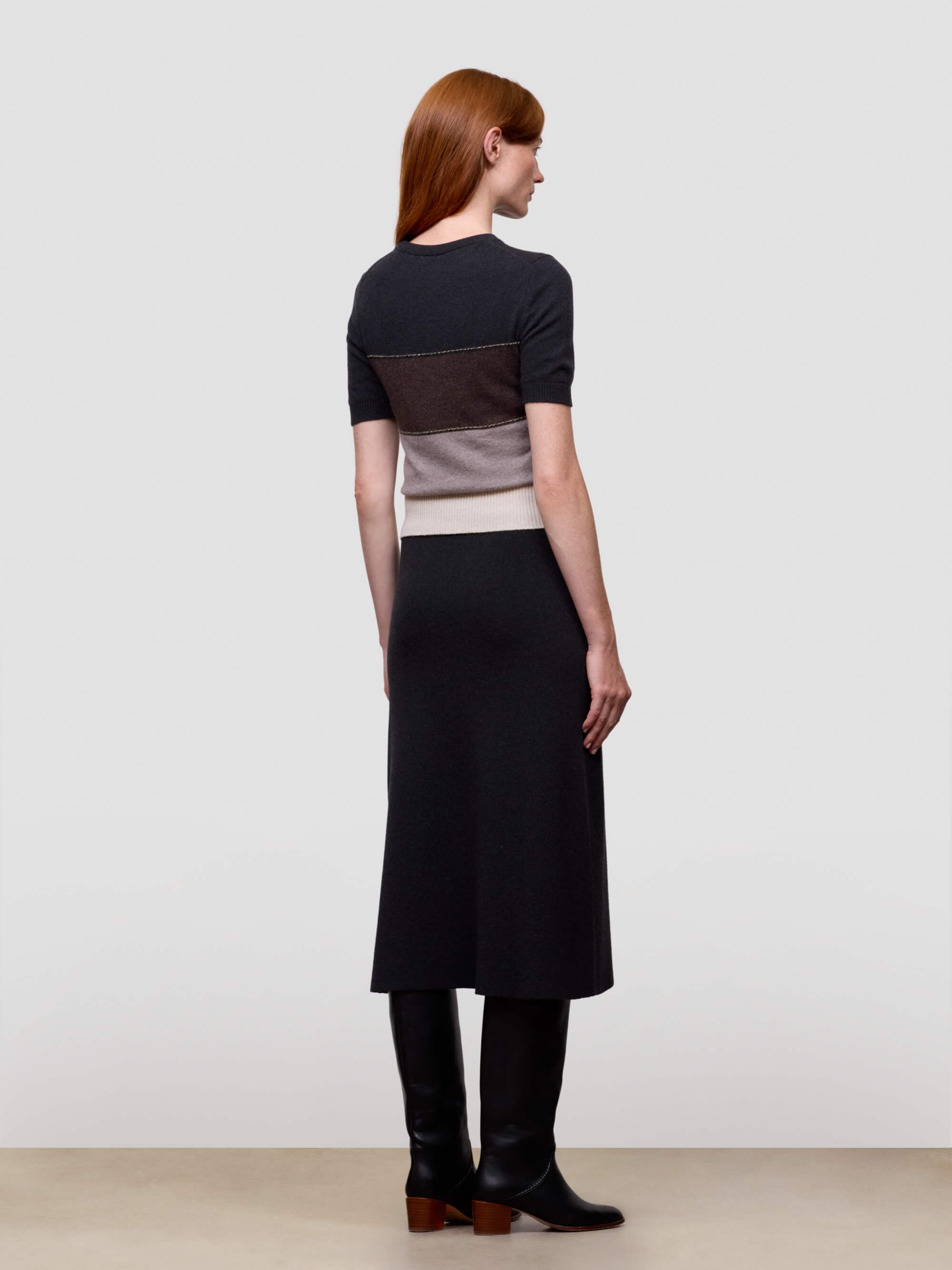 Woman wearing a black, brown, and beige color-blocked cashmere t-shirt on a light gray background and black cashmere dress with boots