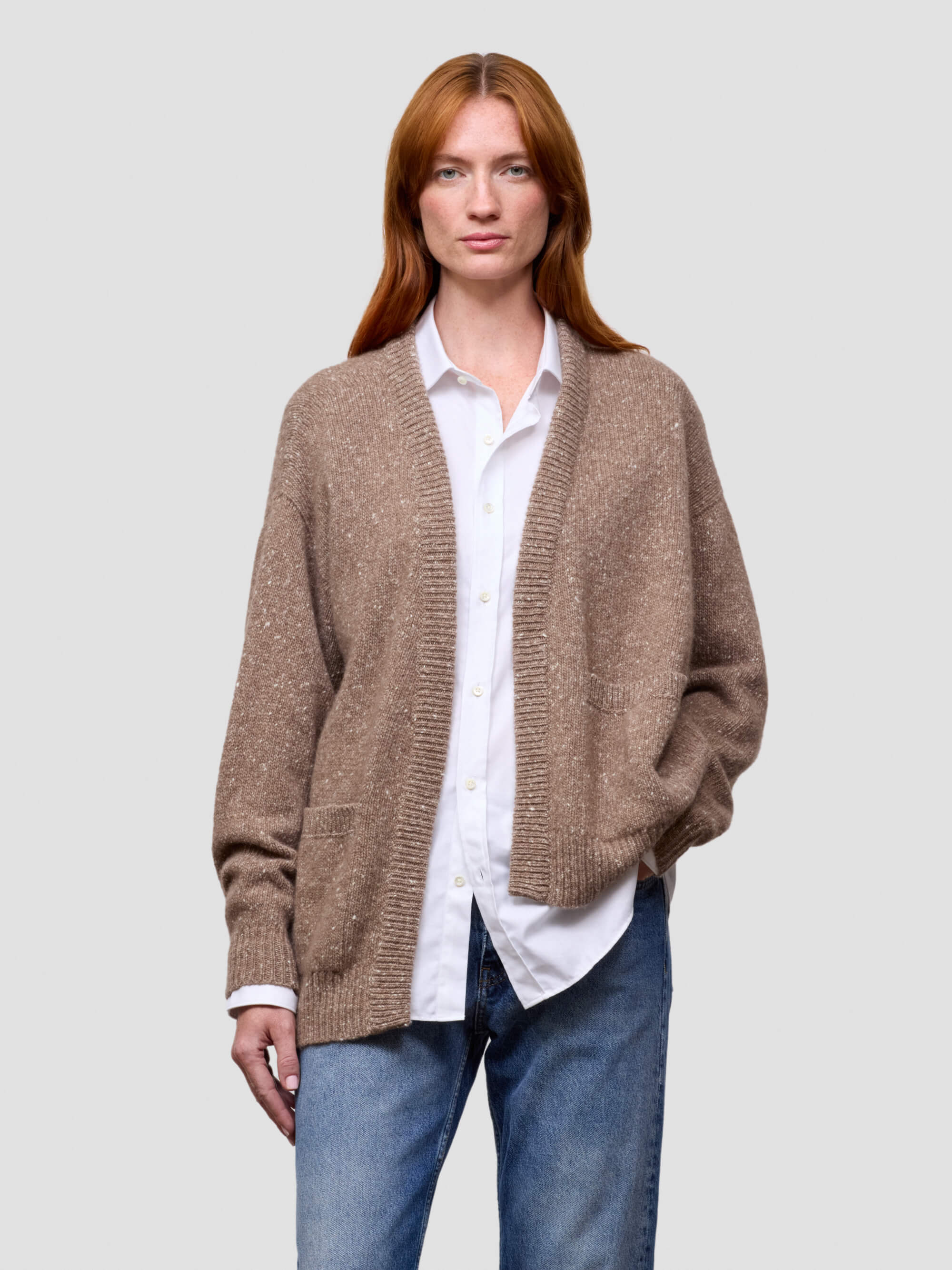 Woman wearing a brown cashmere cardigan over a white shirt and blue jeans on a light gray background