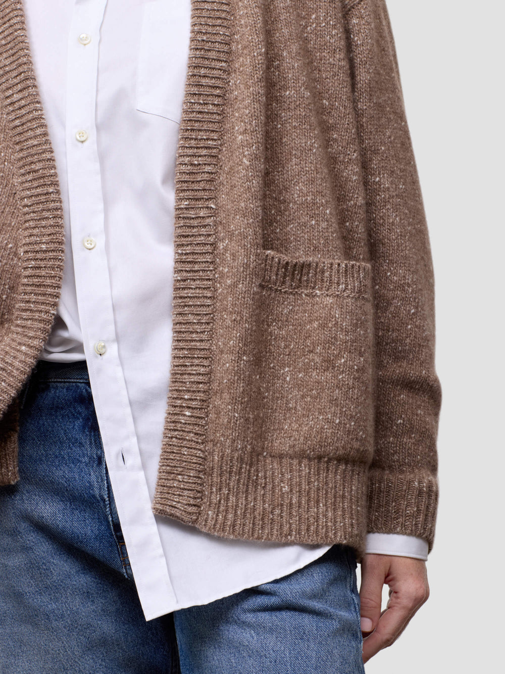 Woman wearing a brown cashmere cardigan over a white shirt and blue jeans on a light gray background