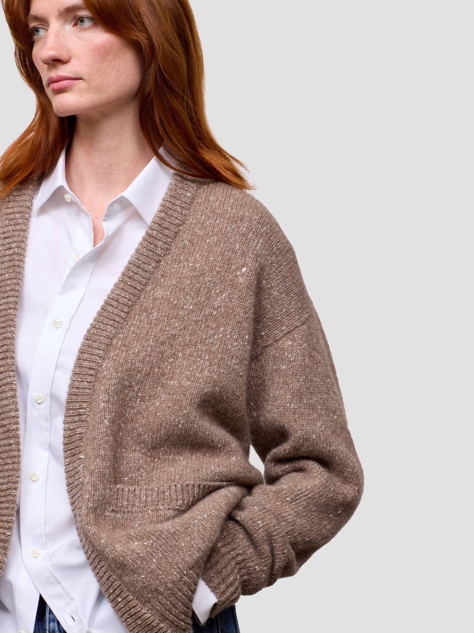 Woman wearing a brown cashmere cardigan over a white shirt and blue jeans on a light gray background