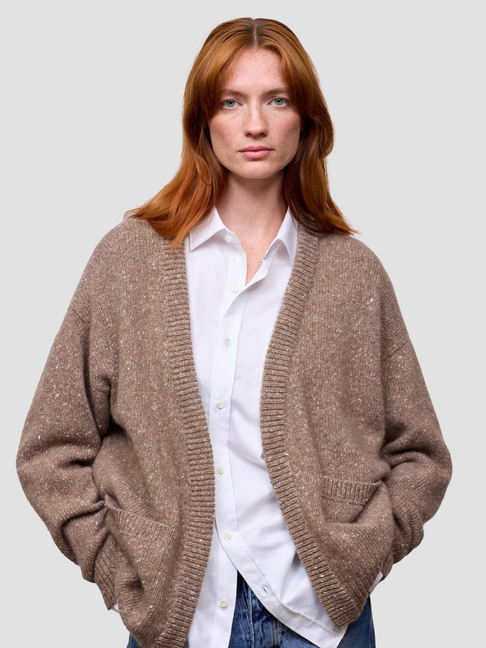 Woman wearing a brown cashmere cardigan over a white shirt and blue jeans on a light gray background