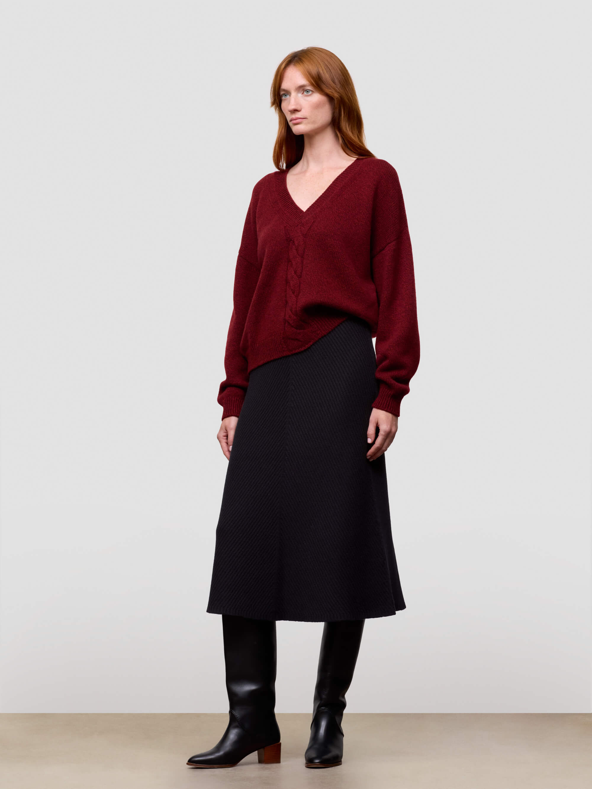 woman wearing a red chunky v-neck cashmere sweater and black cashmere skirt on a grey background