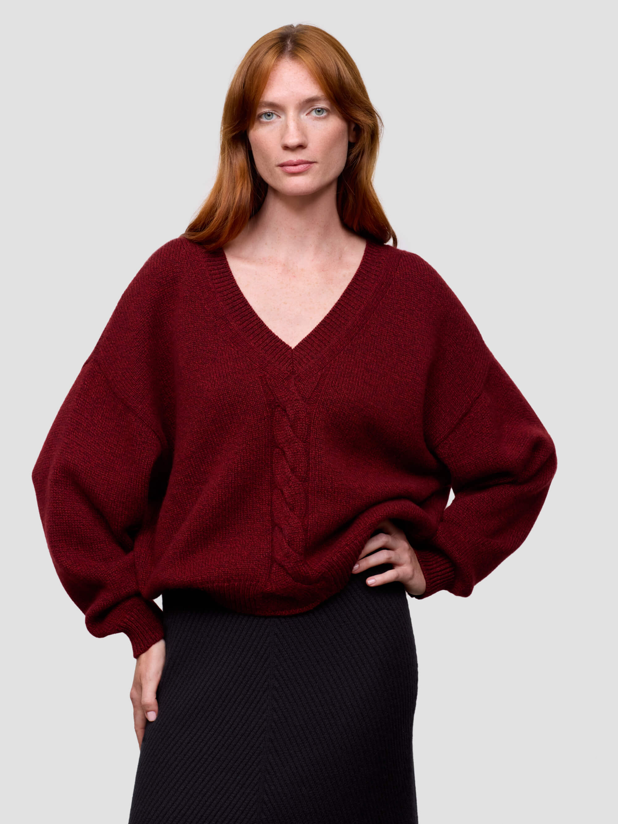 woman wearing a red chunky v-neck cashmere sweater and black cashmere skirt on a grey background