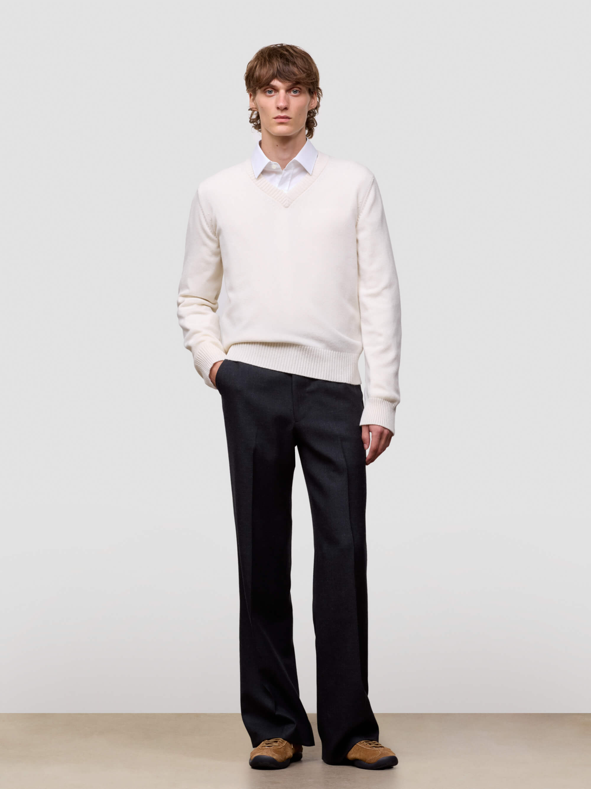 Man wearing a white cashmere v-neck sweater over a collared shirt with dark pants on a light gray background