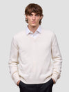 Man wearing a white cashmere v-neck sweater over a collared shirt with dark pants on a light gray background