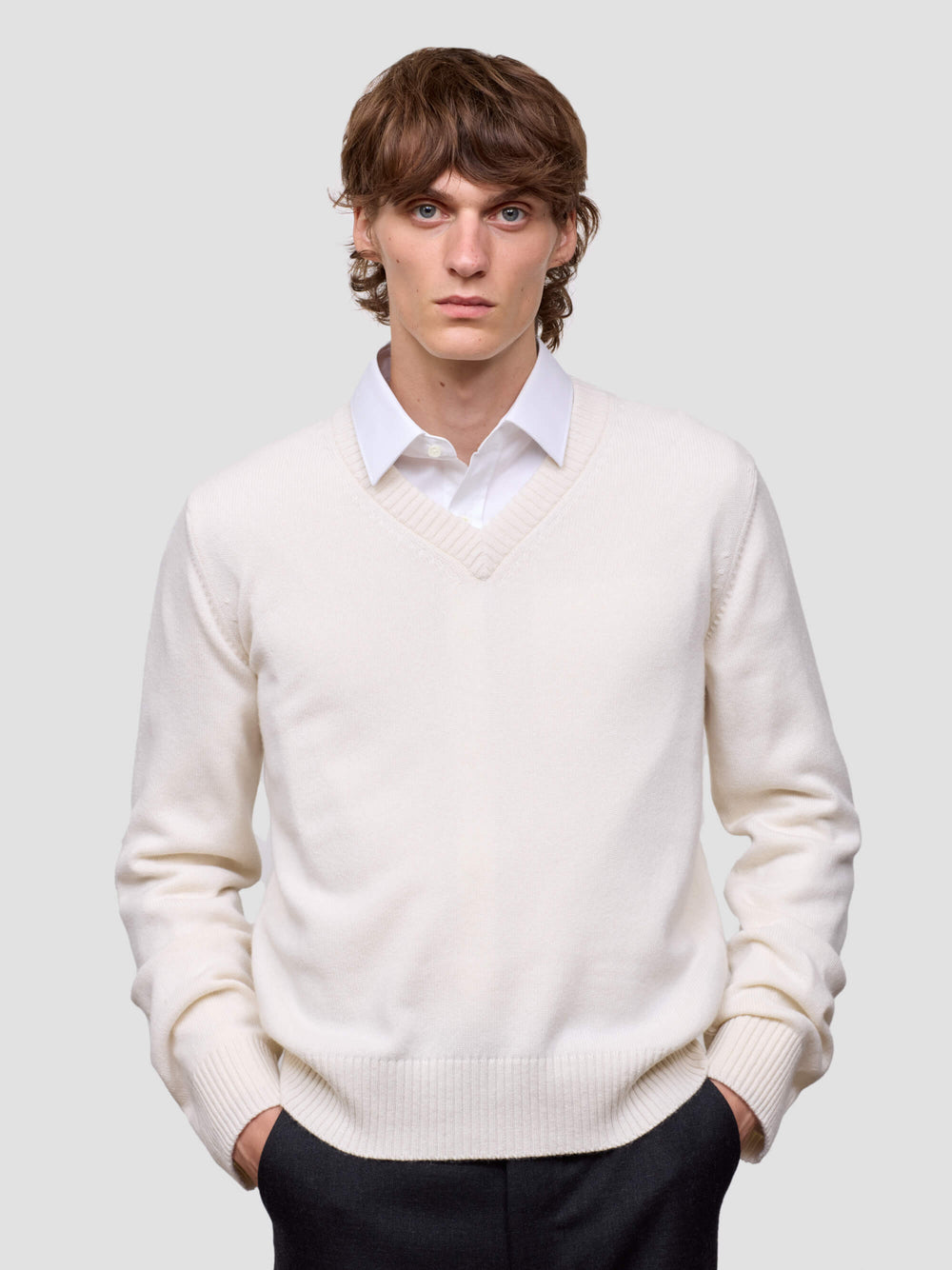 Man wearing a white cashmere v-neck sweater over a collared shirt with dark pants on a light gray background