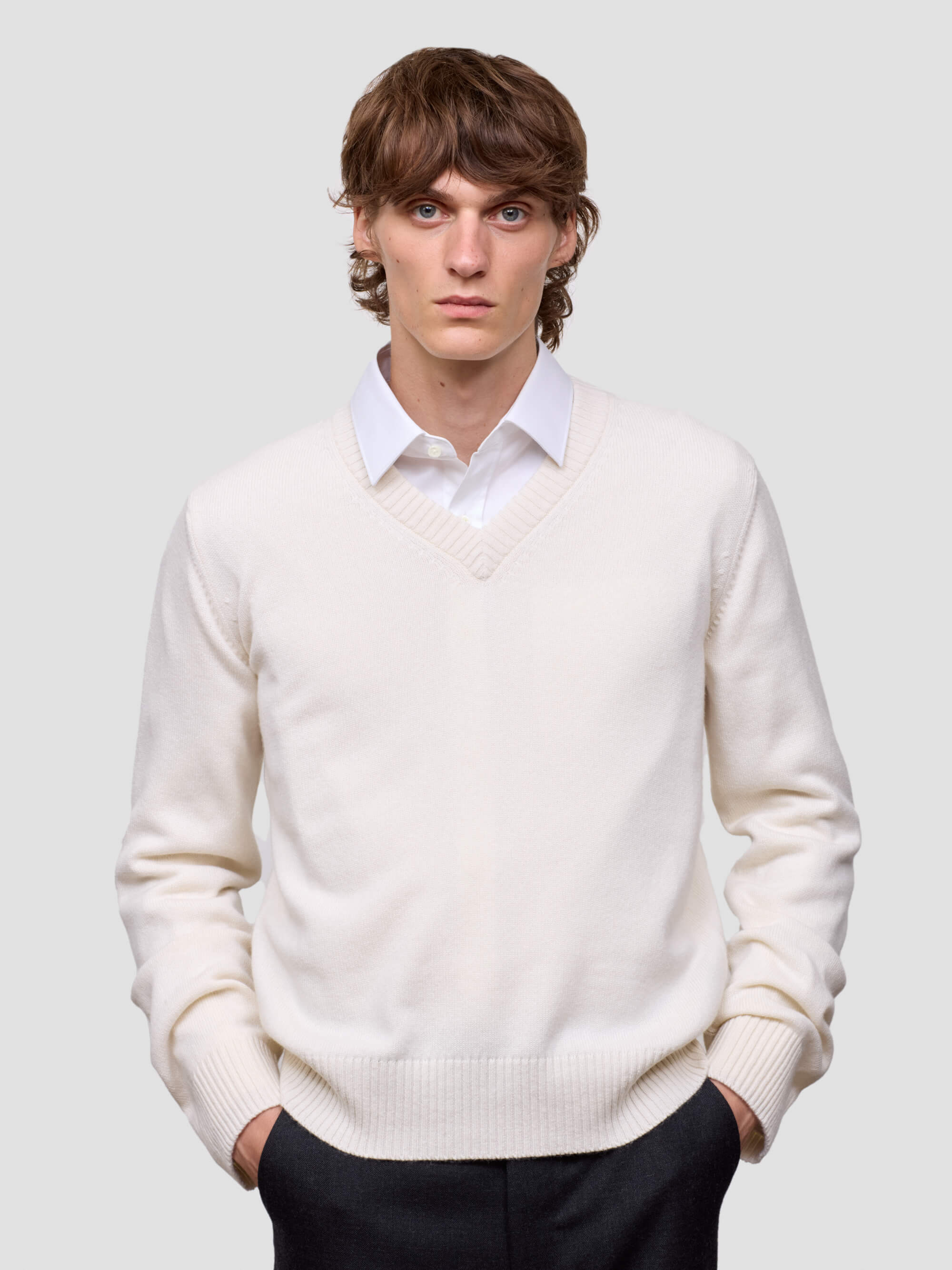 Man wearing a white cashmere v-neck sweater over a collared shirt with dark pants on a light gray background