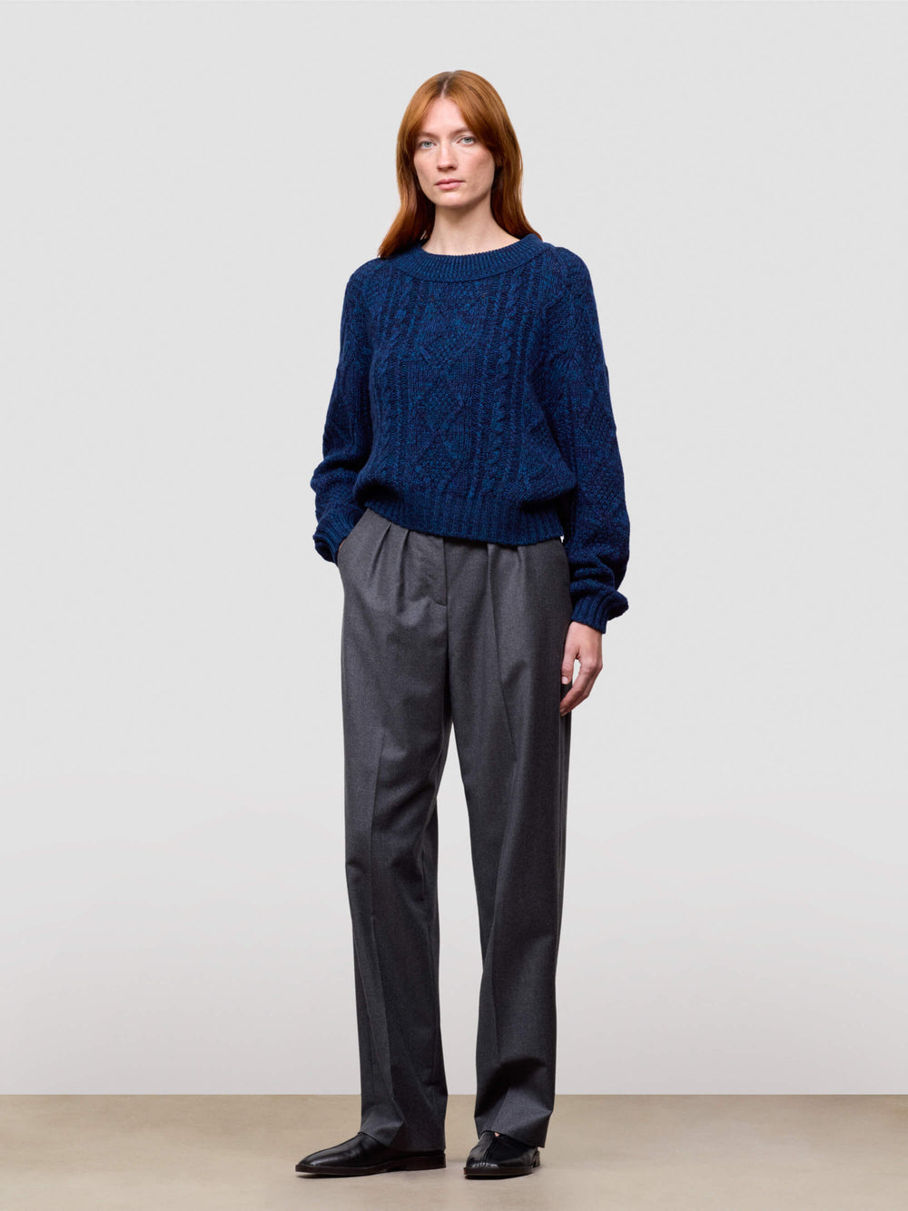 woman wearing a blue cable knit cashmere sweater and gray pants on a plain background