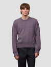 Man wearing a patterned purple cashmere sweater and black pants on a plain background